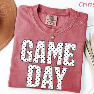 Puede incluir: Una camiseta de manga corta color carmes&iacute; con cuello redondo que presenta el texto GAME DAY en letras grandes de bloque blancas con puntos negros. La camiseta tiene una etiqueta de la marca Comfort Colors en el cuello y es de algod&oacute;n.