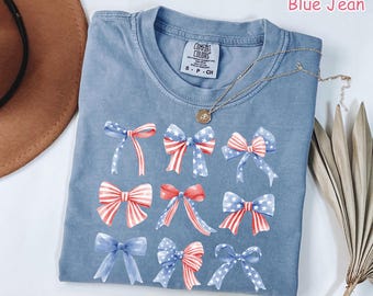 May include: A blue jean colored t-shirt featuring a graphic print of nine distinct bows in various red, white, and blue patterns, including stars and stripes. The shirt has a crew neckline and a Comfort Colors brand label at the inner collar.