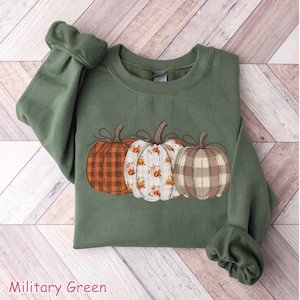Puede incluir: Sudadera verde militar con tres calabazas decorativas con diferentes dise&ntilde;os: cuadros naranjas, floral y cuadros beige. Las calabazas est&aacute;n cosidas en la parte delantera. El texto "Military Green" est&aacute; en la parte inferior.