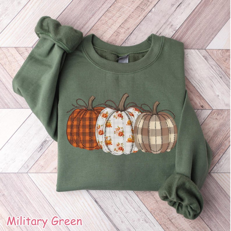 Puede incluir: Sudadera verde militar con tres calabazas decorativas con diferentes dise&ntilde;os: cuadros naranjas, floral y cuadros beige. Las calabazas est&aacute;n cosidas en la parte delantera. El texto "Military Green" est&aacute; en la parte inferior.
