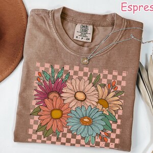 May include: Espresso-colored t-shirt with a floral design. The graphic features colorful daisies on a pink and brown checkered background. A silver necklace with a pendant is draped on the shirt. The shirt is made of a soft material.