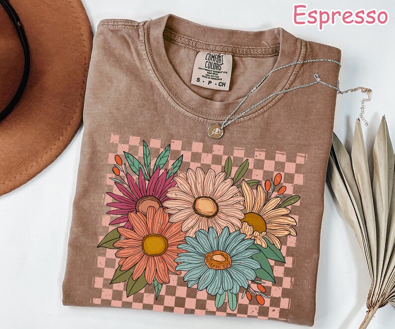 May include: Espresso-colored t-shirt with a floral design. The graphic features colorful daisies on a pink and brown checkered background. A silver necklace with a pendant is draped on the shirt. The shirt is made of a soft material.
