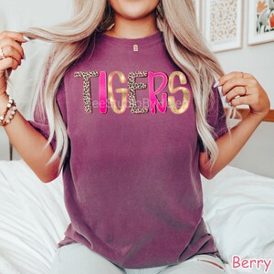 Peut inclure: T-shirt de couleur baie avec le mot "TIGERS" dans un design audacieux et superpos&eacute;. Les lettres ont un contour dor&eacute;, un centre &agrave; imprim&eacute; l&eacute;opard et une ombre rose vif. Le t-shirt est &agrave; manches courtes et &agrave; col rond.