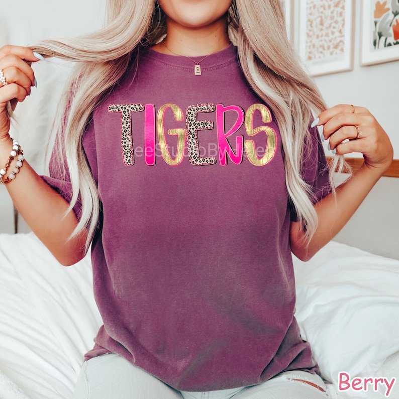 Peut inclure: T-shirt de couleur baie avec le mot "TIGERS" dans un design audacieux et superpos&eacute;. Les lettres ont un contour dor&eacute;, un centre &agrave; imprim&eacute; l&eacute;opard et une ombre rose vif. Le t-shirt est &agrave; manches courtes et &agrave; col rond.