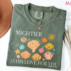 K&ouml;nnte beinhalten: Moosgr&uuml;nes T-Shirt mit dem Text "MIGHTIER than the waves of the sea IS HIS LOVE FOR YOU" und Muschel-Grafiken. Eine silberne Halskette mit Anh&auml;nger liegt auf dem Shirt.