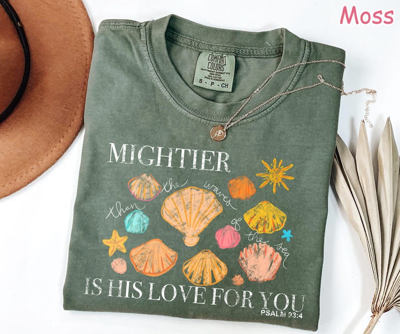 K&ouml;nnte beinhalten: Moosgr&uuml;nes T-Shirt mit dem Text "MIGHTIER than the waves of the sea IS HIS LOVE FOR YOU" und Muschel-Grafiken. Eine silberne Halskette mit Anh&auml;nger liegt auf dem Shirt.