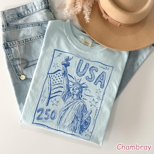Puede incluir: Una camiseta azul claro con un estampado gr&aacute;fico azul oscuro de la Estatua de la Libertad, una bandera estadounidense, estrellas, el texto USA y el n&uacute;mero 250. La prenda se presenta junto a unos vaqueros de mezclilla clara y un sombrero beige.