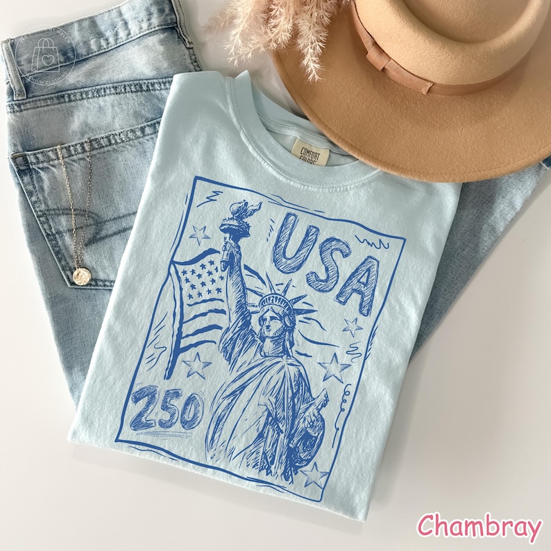 Puede incluir: Una camiseta azul claro con un estampado gr&aacute;fico azul oscuro de la Estatua de la Libertad, una bandera estadounidense, estrellas, el texto USA y el n&uacute;mero 250. La prenda se presenta junto a unos vaqueros de mezclilla clara y un sombrero beige.