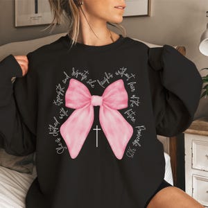 May include: Black sweatshirt with a pink bow and a cross. The text "She is clothed with strength and dignity; she laughs without fear of the future." Proverbs 31:25 is printed on the sweatshirt.