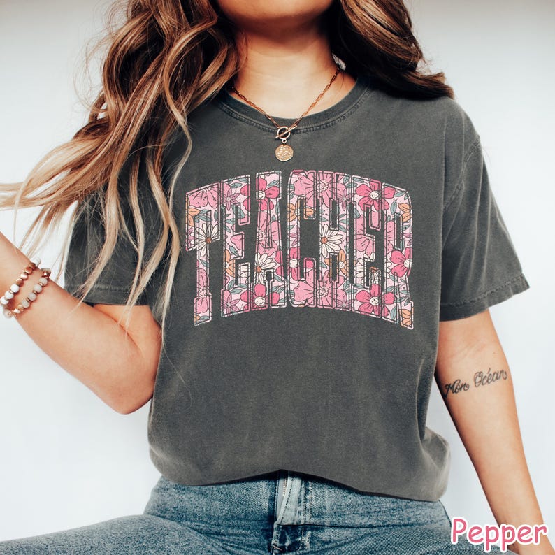 Puede incluir: Una camiseta gris con una impresi&oacute;n floral de la palabra "TEACHER" en rosa y amarillo.