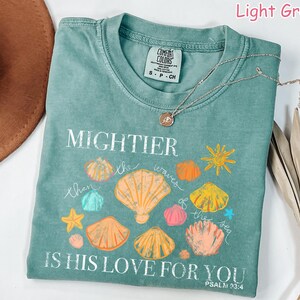 K&ouml;nnte beinhalten: Hellgr&uuml;nes T-Shirt mit dem Text "Mightier than the waves of the sea is His love for you" und Muschel-Grafiken. Das Shirt hat einen Rundhalsausschnitt und kurze &Auml;rmel. Eine silberne Halskette mit Anh&auml;nger befindet sich auf dem Shirt.