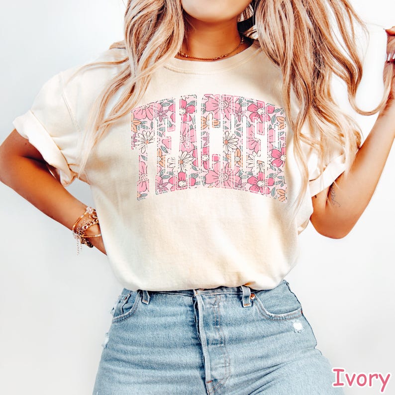 Puede incluir: Una camiseta blanca con un dise&ntilde;o floral que deletrea la palabra "Teacher" en rosa y blanco.
