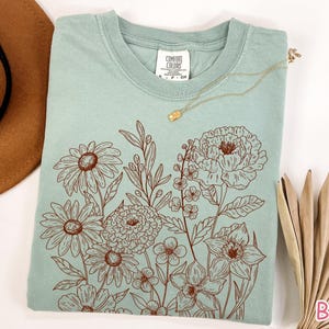 Puede incluir: Una camiseta verde claro con un dise&ntilde;o floral marr&oacute;n. El dise&ntilde;o presenta varias flores y hojas. Un collar dorado con un peque&ntilde;o colgante descansa sobre la camisa. Un sombrero marr&oacute;n y una hoja de palma seca est&aacute;n en el fondo.