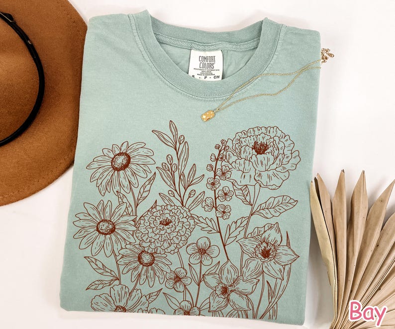 Puede incluir: Una camiseta verde claro con un dise&ntilde;o floral marr&oacute;n. El dise&ntilde;o presenta varias flores y hojas. Un collar dorado con un peque&ntilde;o colgante descansa sobre la camisa. Un sombrero marr&oacute;n y una hoja de palma seca est&aacute;n en el fondo.