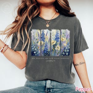 K&ouml;nnte beinhalten: Dunkelgraues T-Shirt mit einem Blumendesign aus blauen, gelben und lila Blumen. Der Text "HIS mercies are new every morning Lamentations 3:23" ist darunter gedruckt. Das T-Shirt hat kurze &Auml;rmel.