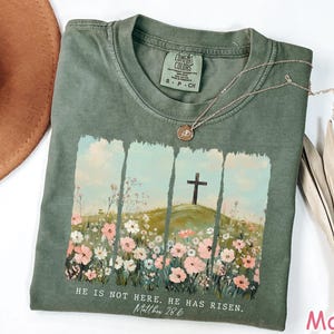 May include: A green t-shirt with a floral design and a cross. The text "HE IS NOT HERE. HE HAS RISEN. Matthew 28:6" is printed on the shirt.