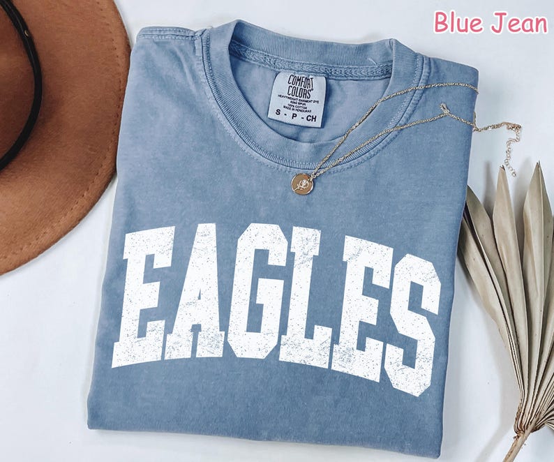 May include: Blue jean Comfort Colors t-shirt with the word "EAGLES" in large white block letters. A gold necklace with a pendant is draped across the shirt. The shirt is folded on a white surface.