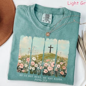 May include: Light green t-shirt with a floral design and a cross. The shirt has the text "HE IS NOT HERE. HE HAS RISEN. Matthew 28.6" printed on the bottom.