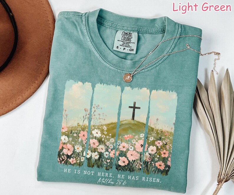 May include: Light green t-shirt with a floral design and a cross. The shirt has the text "HE IS NOT HERE. HE HAS RISEN. Matthew 28.6" printed on the bottom.