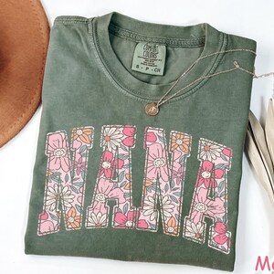 May include: A green t-shirt with a floral design that spells out "NANA". The shirt is made of a soft, comfortable fabric and has a relaxed fit.