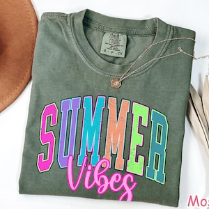 May include: A moss green t-shirt with the words "SUMMER VIBES" in a colorful, outlined font. A gold necklace with a pendant rests on the shirt. A brown hat and dried palm leaves are visible.
