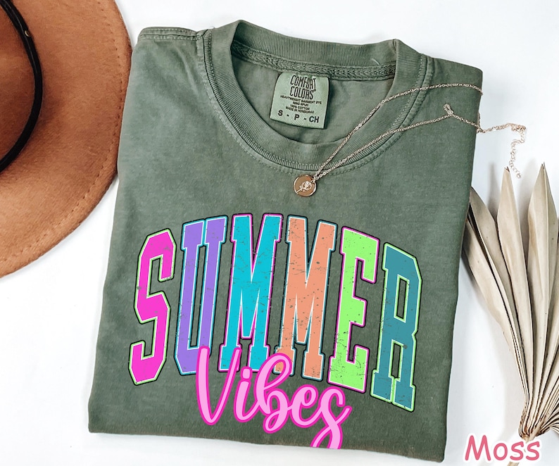 May include: A moss green t-shirt with the words "SUMMER VIBES" in a colorful, outlined font. A gold necklace with a pendant rests on the shirt. A brown hat and dried palm leaves are visible.