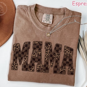 May include: Espresso-colored t-shirt with the word "MAMA" in large, bold letters. The letters alternate between a checkered pattern and a leopard print design. A silver necklace with a small pendant rests on the shirt.