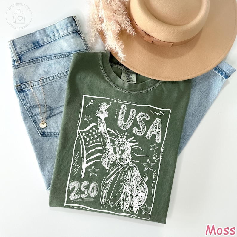Puede incluir: Una camiseta verde musgo con un gr&aacute;fico de l&iacute;neas blancas de la Estatua de la Libertad, una bandera estadounidense, el texto USA y el n&uacute;mero 250. La prenda se presenta sobre unos vaqueros azul claro, un sombrero beige y una fina cadena dorada.