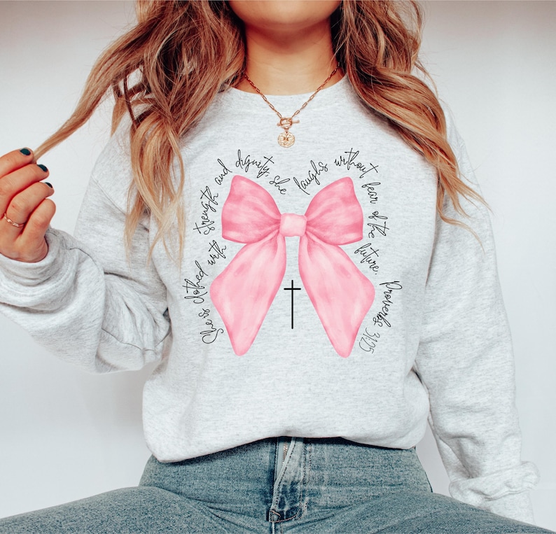 May include: A gray sweatshirt with a pink bow and the text "She is clothed with strength and dignity, she laughs without fear of the future. Proverbs 31:25"