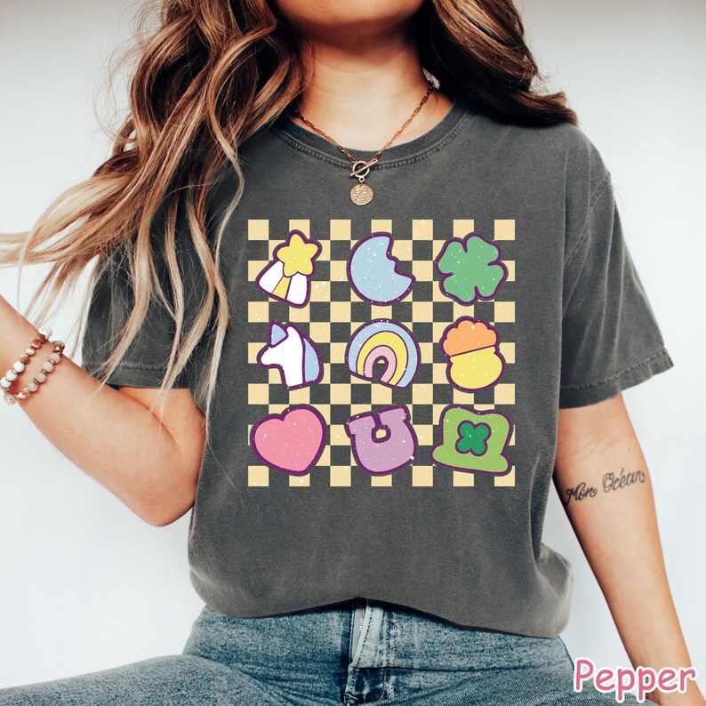Peut inclure: Un t-shirt gris &agrave; motif &agrave; carreaux avec une vari&eacute;t&eacute; d'images de dessins anim&eacute;s color&eacute;s, notamment un arc-en-ciel, un fer &agrave; cheval, un c&oelig;ur, un tr&egrave;fle &agrave; quatre feuilles, une licorne, un pot d'or et une &eacute;toile. Le texte "No Drama" est &eacute;crit sur la manche droite du t-shirt.