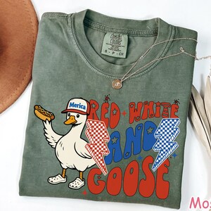 May include: A moss green t-shirt with a graphic of a goose wearing a "Merica" hat and holding a hot dog. The shirt features the text "Red White and Goose" in red and blue, with lightning bolt designs. A gold necklace is draped on the shirt.