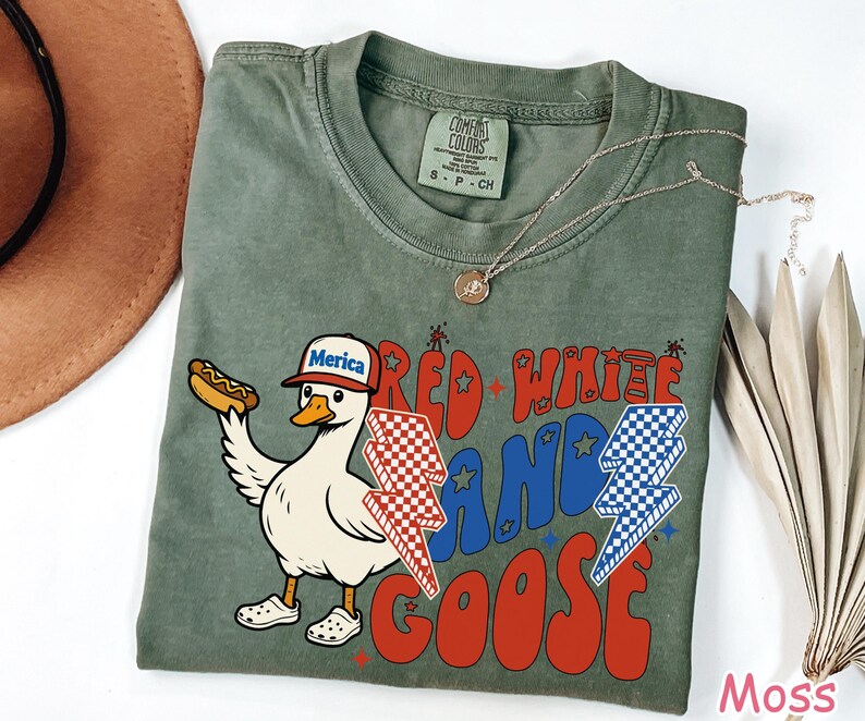May include: A moss green t-shirt with a graphic of a goose wearing a "Merica" hat and holding a hot dog. The shirt features the text "Red White and Goose" in red and blue, with lightning bolt designs. A gold necklace is draped on the shirt.