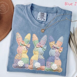 May include: Blue jean Comfort Colors t-shirt with three appliqued patchwork bunny rabbits. Each bunny has a bow and a white flower. A gold necklace is draped across the shirt. The text "Blue Jean" is in the upper right corner.