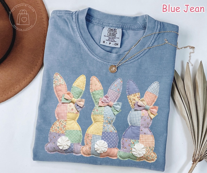 K&ouml;nnte beinhalten: Blaues Jeans Comfort Colors T-Shirt mit drei aufgen&auml;hten Patchwork-Hasen. Jeder Hase hat eine Schleife und eine wei&szlig;e Blume. Eine goldene Halskette liegt &uuml;ber dem Shirt. Der Text "Blue Jean" befindet sich in der oberen rechten Ecke.