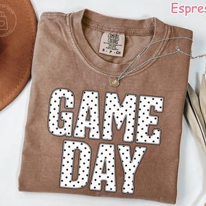Puede incluir: Una camiseta de manga corta en color marr&oacute;n espresso con cuello redondo. En la parte delantera destaca el texto GAME DAY en letras grandes y blancas con un patr&oacute;n de puntos negros. Un collar plateado con un peque&ntilde;o colgante descansa sobre la prenda.