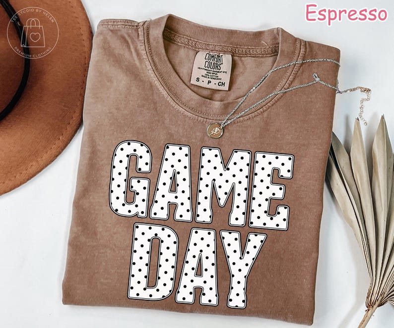 Puede incluir: Una camiseta de manga corta en color marr&oacute;n espresso con cuello redondo. En la parte delantera destaca el texto GAME DAY en letras grandes y blancas con un patr&oacute;n de puntos negros. Un collar plateado con un peque&ntilde;o colgante descansa sobre la prenda.
