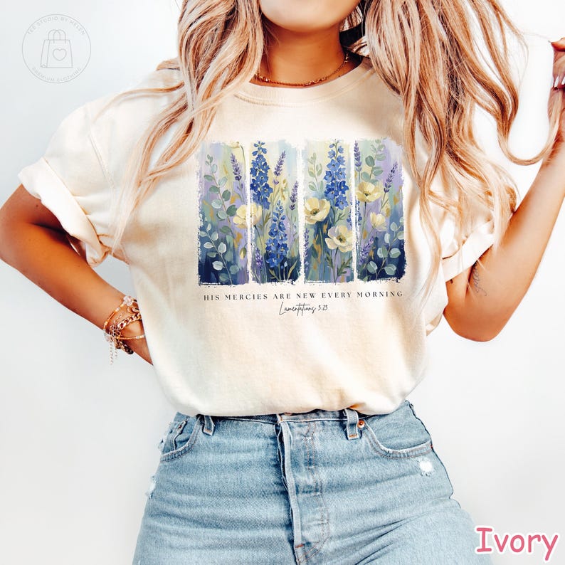 K&ouml;nnte beinhalten: Elfenbeinfarbenes T-Shirt mit einem Blumendesign mit blauen, gelben und gr&uuml;nen Blumen. Der Text "HIS MERCIES ARE NEW EVERY MORNING" ist unter dem Blumendesign aufgedruckt, darunter "Lamentations 3:23".