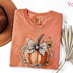 May include: A coral-colored Comfort Colors t-shirt featuring a pumpkin design with a polka-dot bow and floral accents. A gold necklace and a brown hat are next to the shirt. The word "Yam" is in the upper right corner.