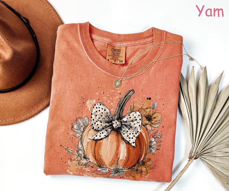 May include: A coral-colored Comfort Colors t-shirt featuring a pumpkin design with a polka-dot bow and floral accents. A gold necklace and a brown hat are next to the shirt. The word "Yam" is in the upper right corner.