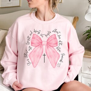 May include: Pink sweatshirt with a large pink bow and a cross. The text on the sweatshirt reads "She is clothed with strength and dignity. She laughs without fear of the future. Proverbs 31:25."