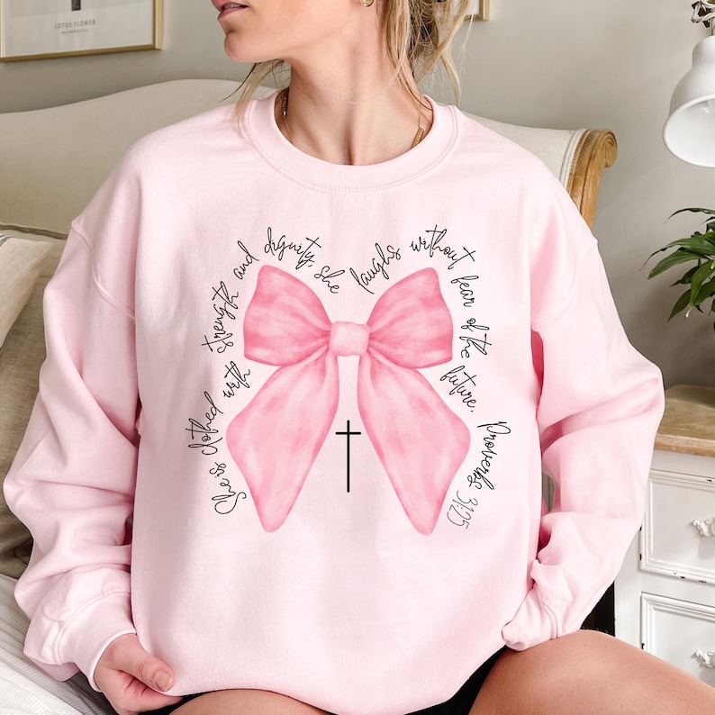 May include: Pink sweatshirt with a large pink bow and a cross. The text on the sweatshirt reads "She is clothed with strength and dignity. She laughs without fear of the future. Proverbs 31:25."