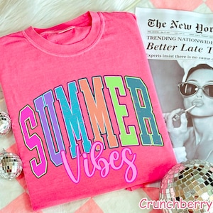 May include: A bright pink t-shirt with the words "SUMMER Vibes" in a colorful, collegiate-style font. The shirt is displayed with a newspaper and disco balls, creating a fun, summery vibe.