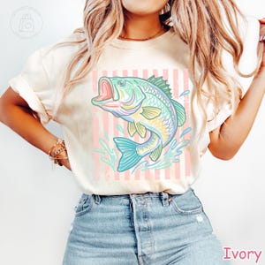 May include: An ivory short-sleeve t-shirt featuring a graphic print of a jumping fish in shades of green, yellow, and blue. The fish is positioned over a background of vertical light pink stripes, with blue water splashes surrounding the fish.
