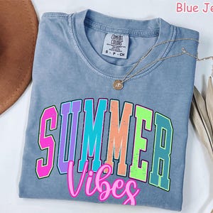 May include: Blue jean colored t-shirt with the words "SUMMER Vibes" in a colorful, collegiate-style font. A gold necklace with a pendant rests on the shirt. The shirt is folded on a white surface.