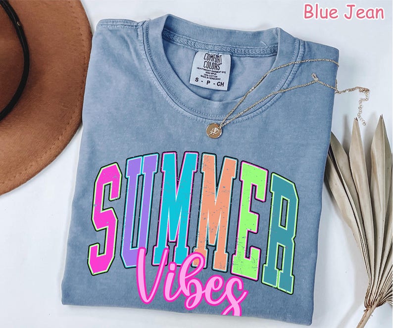 May include: Blue jean colored t-shirt with the words "SUMMER Vibes" in a colorful, collegiate-style font. A gold necklace with a pendant rests on the shirt. The shirt is folded on a white surface.