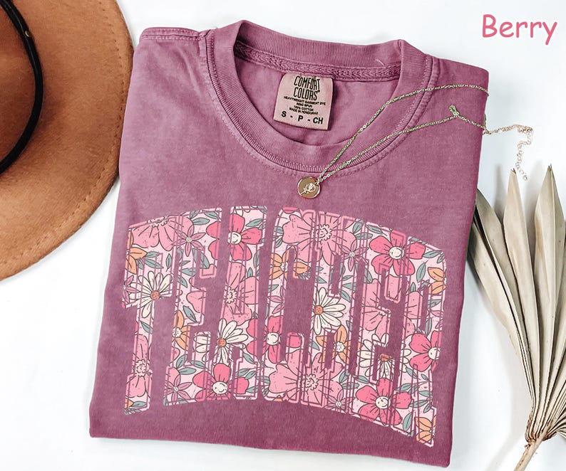 Puede incluir: Una camiseta de color malva con un dise&ntilde;o de estampado floral que deletrea "Teacher" en una fuente de estilo vintage. La camiseta est&aacute; hecha por Comfort Colors y es talla S-P-CH.