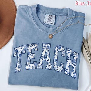 May include: A light blue t-shirt with the word "TEACH" printed in white with a blue and white floral pattern. The shirt is made of a soft, comfortable material.