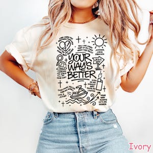 Op de afbeelding: Ivoor t-shirt met een zwart grafisch ontwerp. Het ontwerp bevat tekst en illustraties van een hart, zon, jetski en andere symbolen. De tekst bevat de zin "Your Way's Better."