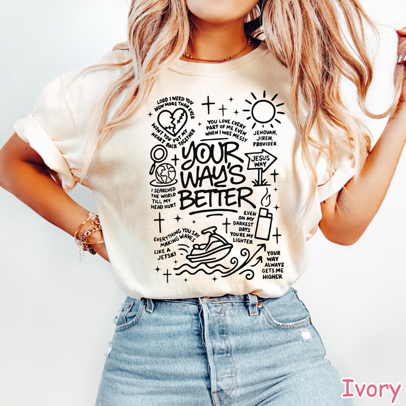 Op de afbeelding: Ivoor t-shirt met een zwart grafisch ontwerp. Het ontwerp bevat tekst en illustraties van een hart, zon, jetski en andere symbolen. De tekst bevat de zin "Your Way's Better."