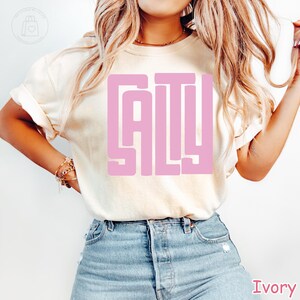 May include: Ivory-colored t-shirt with the word "SALY" in a pink, block letter design. The shirt is made of a soft material and is paired with blue denim jeans. The shirt is a casual style.